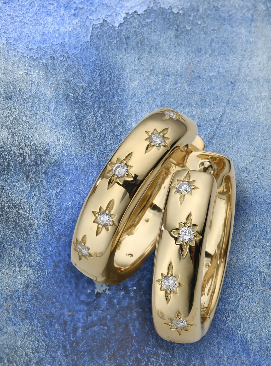a pair of 18k gold huggie hoops set with diamond stars, on a blue stone surface